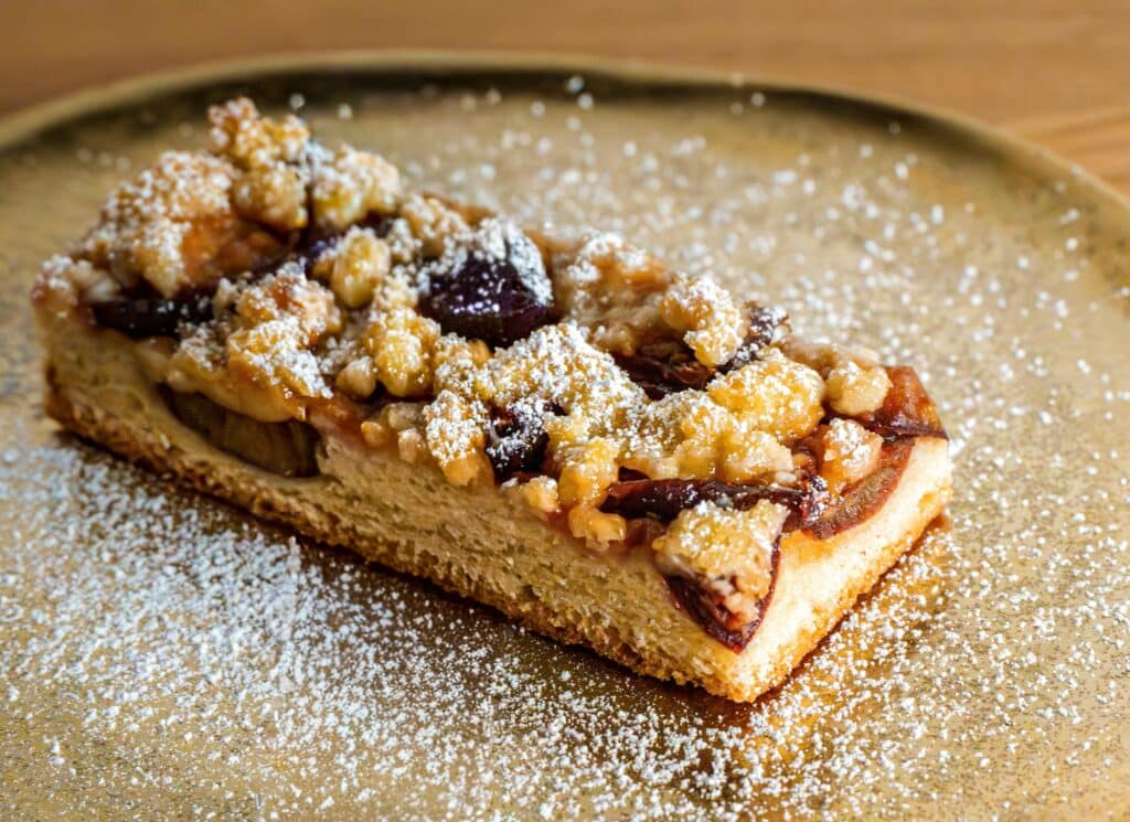 Stück Pflaumenkuchen mit Streuseln und Puderzucker auf goldenem Teller – herbstliche Inspiration für den Duft der AVA & MAY PLUM CAKE Duftkerze.